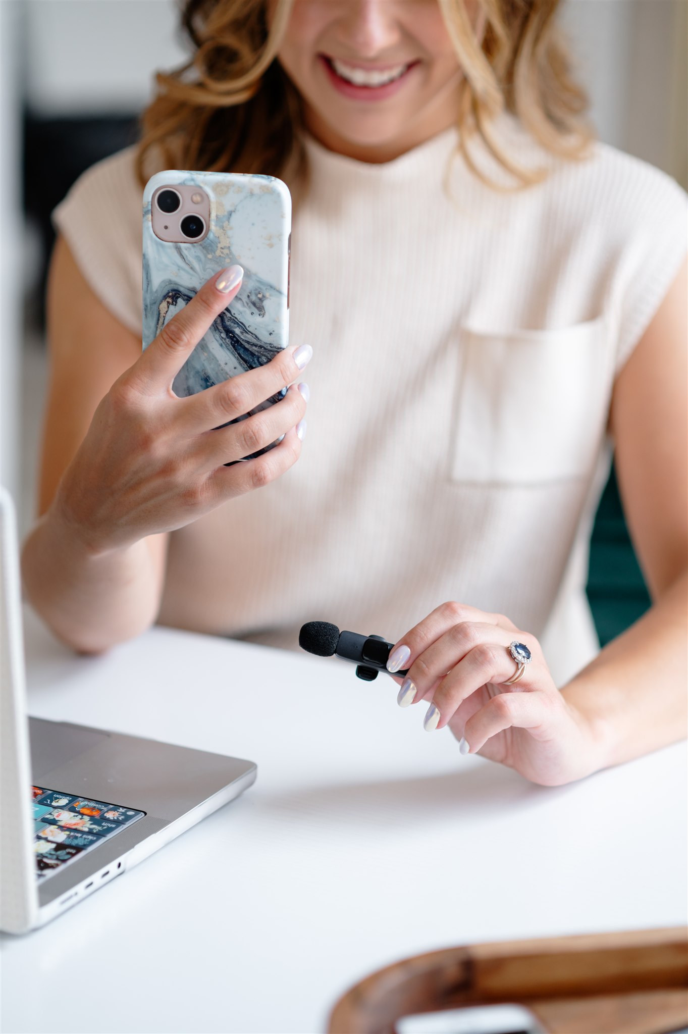 Bee Santangelo, a smiling Massachusetts social media strategist, holds a smartphone and lavalier microphone while sitting at a desk with an open laptop, preparing to record content for social media marketing.