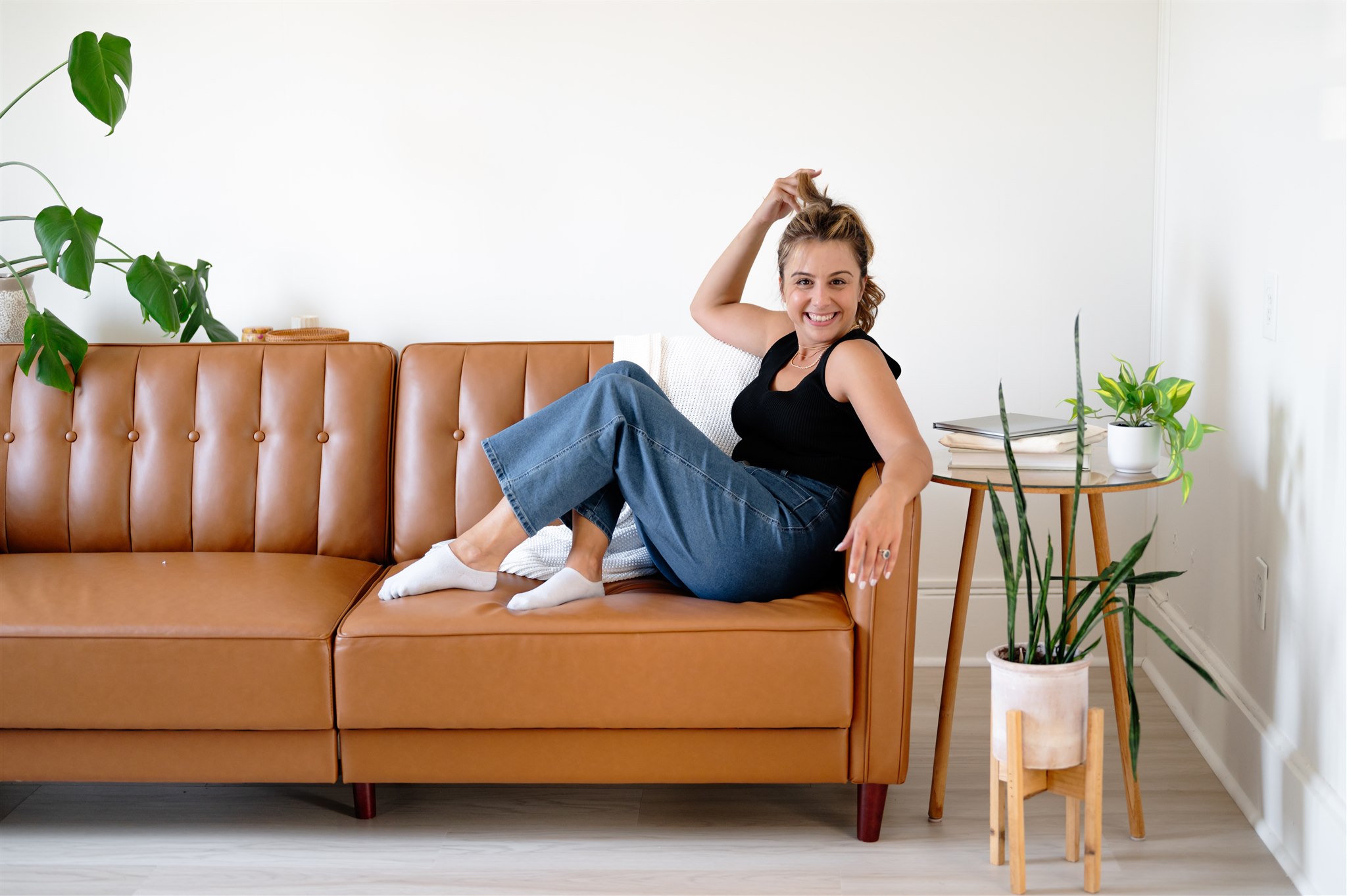 Bee Santangelo, Massachusetts social media manager, lounges on a brown leather sofa, smiling, with plants and books on a side table.